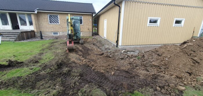 Minigrävare vid garage på gräsmatta med jord och grus, visar pågående markarbete och stenarbeten runt huset.