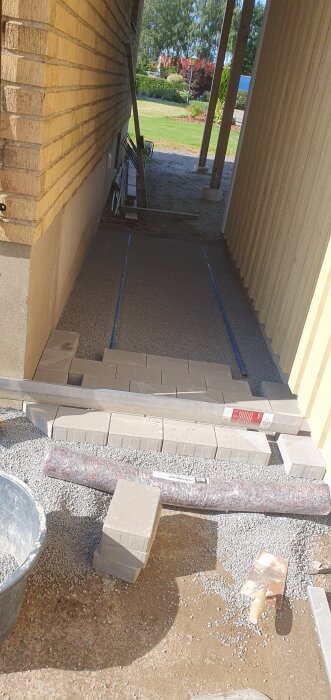 Stenläggning och murbygge vid ett garage, med kantsten och grus runt om. verktyg och material syns i bild.
