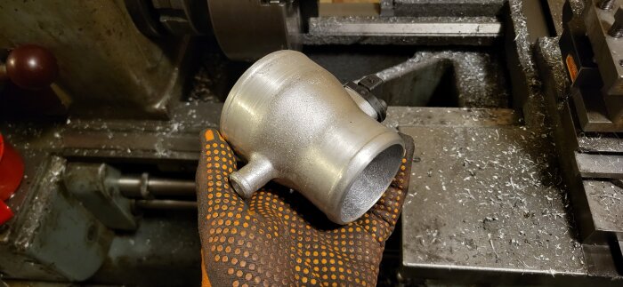 En hand i en prickig handske håller en metallkomponent framför en svarv i en verkstadsmiljö.