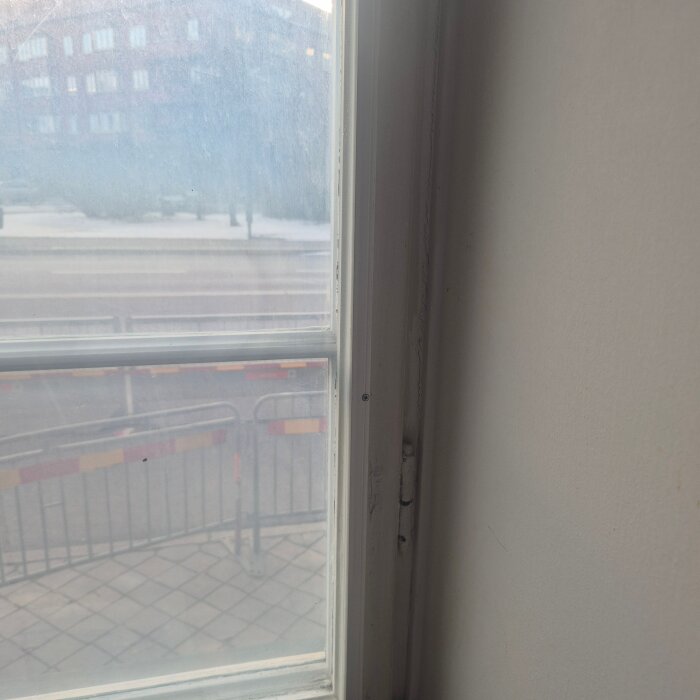 Närbild på gammalt fönster med glipa och utsikt mot en gata. Byggnadsställning och trottoar syns utanför fönstret.