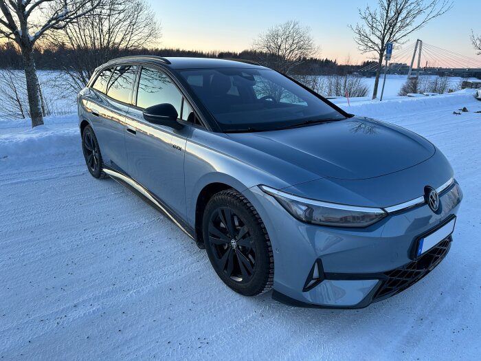 En nytvättad grå bil parkerad på en snötäckt väg med en bro och träd i bakgrunden under solnedgången.