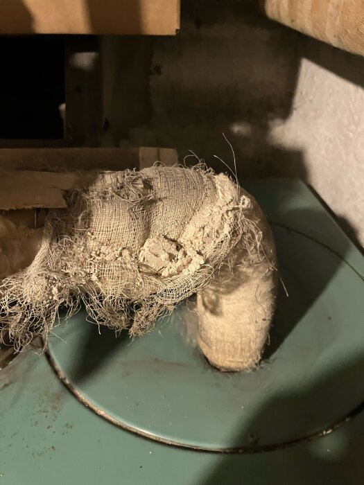 Isolerat gammalt rör med trasigt material, delvis exponerad yta, omgiven av betongvägg och annan isolering i dunkelt utrymme.