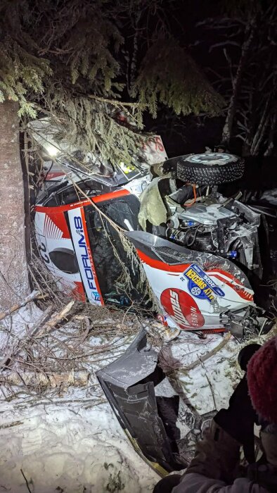 Rallybil vält vid snötäckt väg, träd och grenar nära. Delar av bilen skadade, nattfoto.