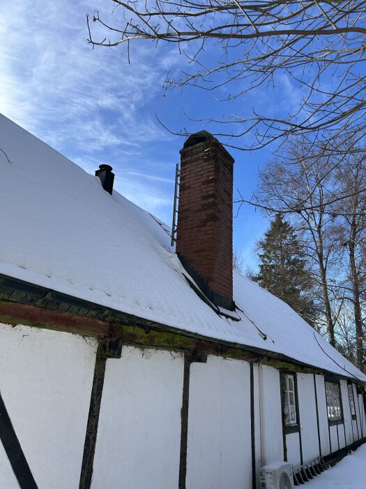 Et gammelt hus med hvite vegger og sort bindingsverk, snødekket tak, synlig skorstein av murstein, omgitt av trær og klar blå himmel.