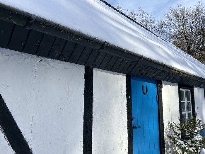 Hvitt hus med blå dør og svarte detaljer under et snødekt tak. En hestesko henger over døren, og det er et tre ved siden av huset.