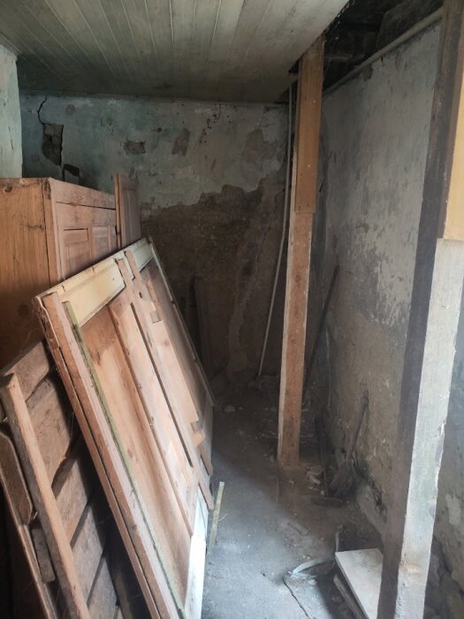 En gammal ekonomibyggnad med slitna väggar och tak, träpaneler lutade mot en vägg och tecken på erosion och förfall.