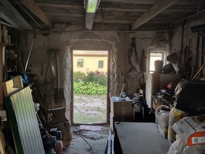En verkstad med verktyg och material, sett genom en dörröppning mot en blomstrande trädgård och ett hus i bakgrunden.