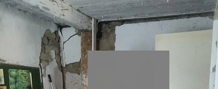 Väggar med synliga sprickor och skador i ett husrum, exponerat trä och puts som lossnat från betongen. Snickarverktyg syns i förgrunden.
