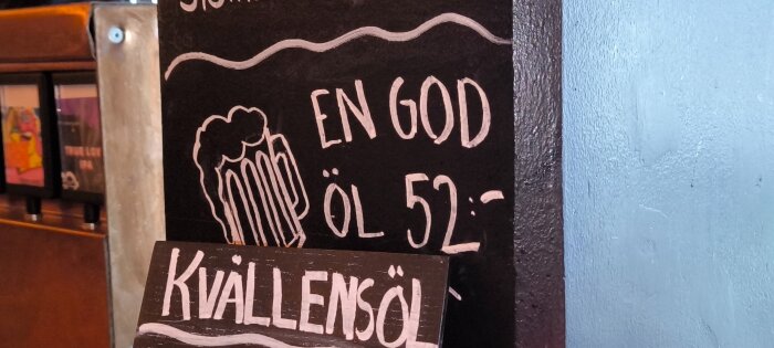Svart tavla med texten "En god öl 52:-" och "Kvällens öl" samt tecknad bild av ett ölglas. Tavlan står mot en blå vägg.