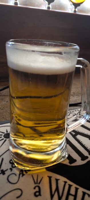Ett glas fyllt med gyllene öl, toppat med skum, står på ett bord med ett dekorativt underlägg.