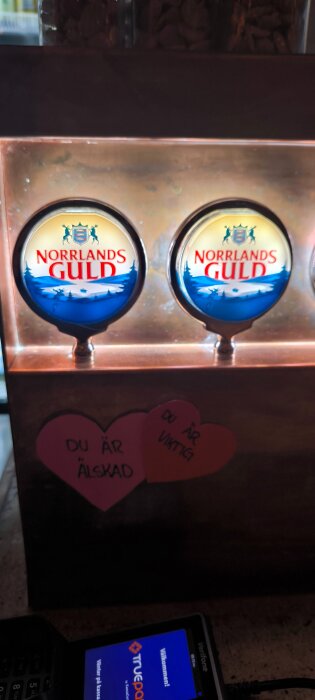 Ölkranar med logotypen för Norrlands Guld. Två hjärtformade lappar med texten "Du är älskad" och "Du är viktig" finns nedanför.
