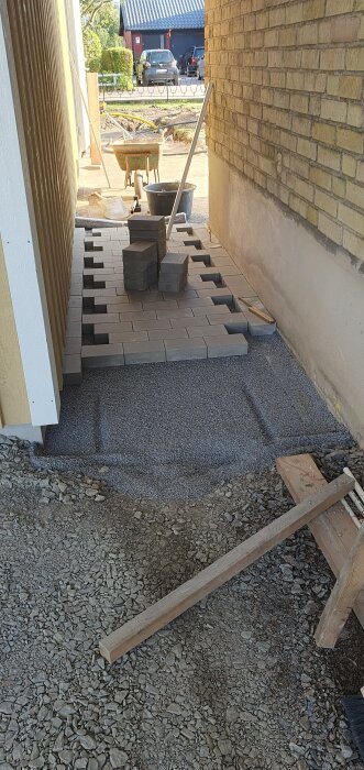 Ny stenläggning påbörjad vid garage, staplade grå stenar, grusgrund och en skottkärra i bakgrunden. Verktyg och material synliga.
