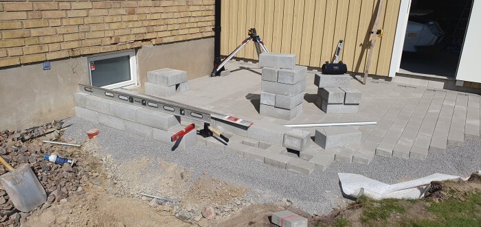 Byggprojekt vid garage med staplade betongblock, verktyg som vattenpass och hammare synliga, samt markarbete med flis och stenläggning påbörjad.
