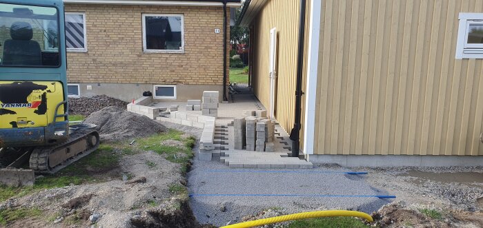 Minigrävare vid pågående stenläggning runt garage med staplade murblock och grus.