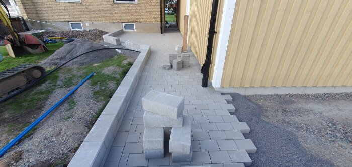 Bild av en nylagd stengång med lösa betongblock vid sidan, intill en gul vägg med påbörjad mur vid ett garage.