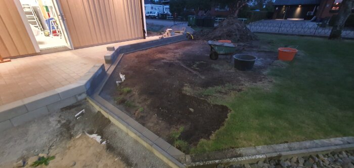 Byggprojekt med ny stenmur och gräsyta kring garage, inklusive schaktjord, flis och arbetsmaterial som skottkärra och hinkar.