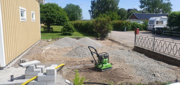 En byggarbetsplats med en padda och grushögar, gjuten betongmur och platta, samt ett beige hus och en husvagn i bakgrunden.