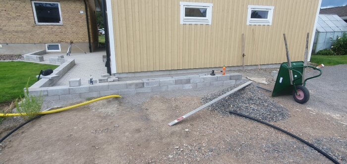 Gjutna cementblock vid gult hus; en mur är uppbyggd bredvid entrén, med staplade block och en skottkärra i förgrunden.