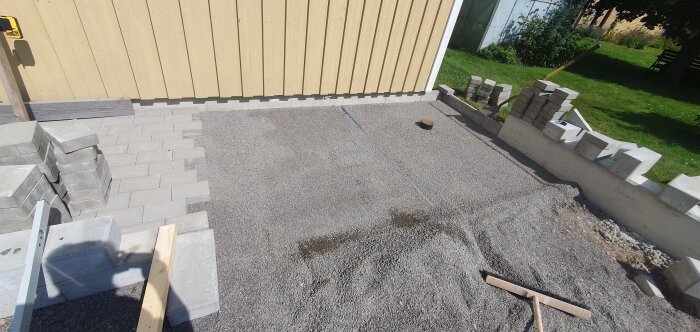 Grusplan framför ett gult trähus under konstruktion med staplade stenplattor, en betongmur och verktyg synliga på bilden.