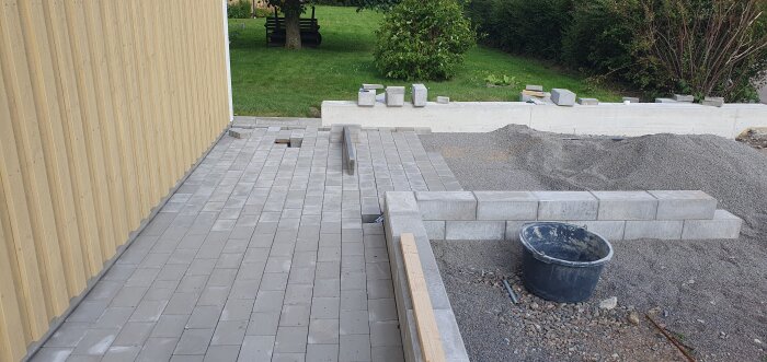 Nyfogat stengolv bredvid gul vägg, staplade betongblock, en betongmur och en svart hink i förgrunden, samt gräsmatta i bakgrunden.