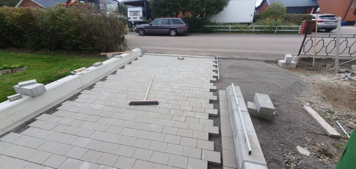 Nybelagd stengång med betongblock längs kanten, en kvast och andra redskap synliga; trädgård och bilar i bakgrunden.