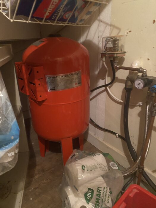Röd hydropress i en skrubb med trägolv, omgiven av rör och emballerade papper.