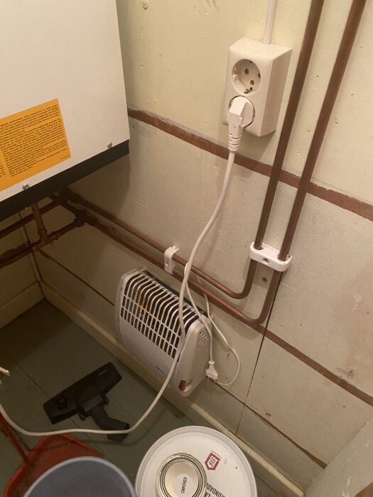 Skrubb med trägolv i en sommarstuga, innehållande varmvattenberedare, hydropress, elektrisk uttag, ledningar, och diverse föremål som hink och dammsugarmunstycke.