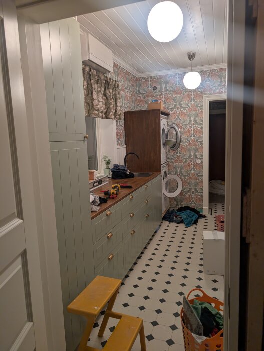 Tvättstuga under renovering med gröna skåpluckor, svartvit rutigt golv, och vit takpanel. Stege, tvättmaskin och torktumlare syns i bilden.