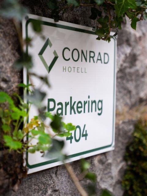 Skylt för "Conrad Hotell Parkering 404" monterad på en stenmur, omgiven av växter.