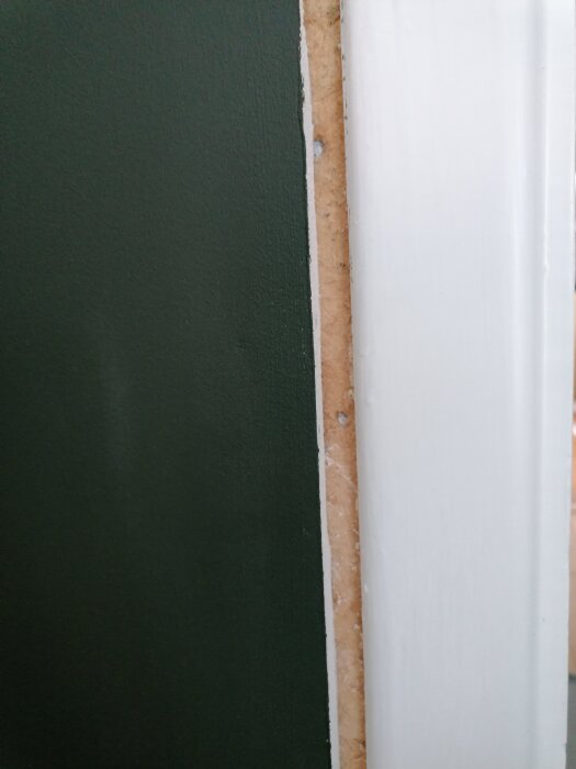 Närbild av en vägg med en mörkgrön målad yta intill en vitmålad dörrkarm, med en smal remsa av osminkad yta synlig emellan.