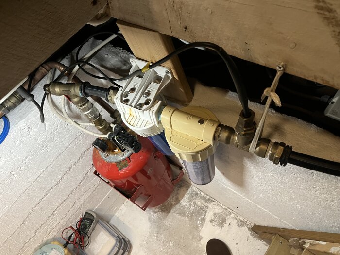 Grovkopplat VVS-system med hydropress, filter och kopplingar av varierande kvalitet under ett golv, med en röd tank närmast väggen.