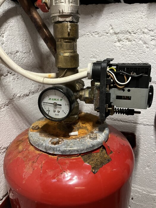 Sliten hydropress med tryckmätare och kablar, röd tank med rost, placerad mot en vit vägg i ett vattenanslutningssystem.