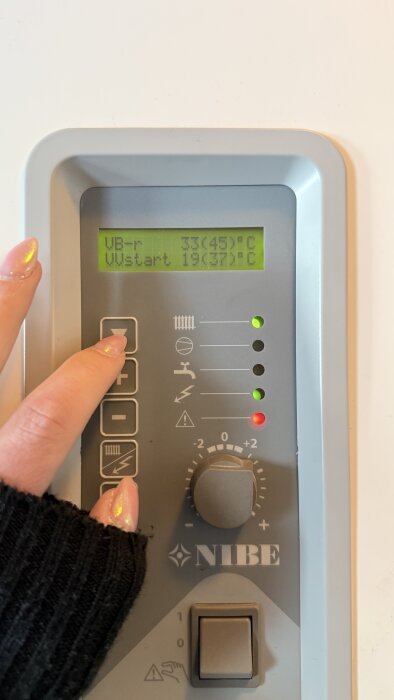 Styrpanel på en värmepump med display som visar temperatur och knappar, person trycker på en knapp. Märket NIBE syns tydligt på panelen.