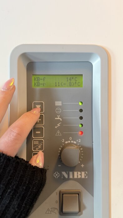 Instrumentpanel på värmepump med temperaturer, en hand trycker på en knapp. Temperaturerna visar "KB-f 14°C" och "KB-r 11(-8)°C".