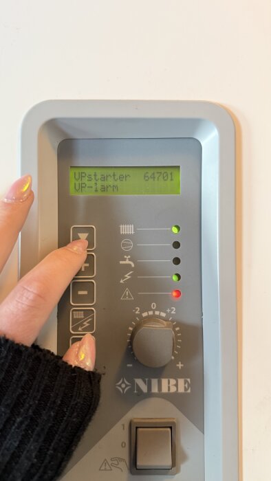 En hand trycker på en knapp på en NIBE-pumpdisplay som visar "VPstarter 64701 VP-larm", med lysdioder som indikerar status.