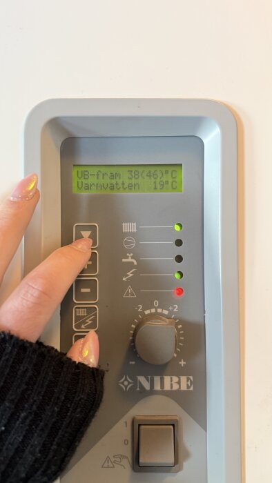 Närbild av NIBE-värmepumps display som visar temp: VB-fram 38(46)°C, Varmvatten 19°C. En hand trycker på knapparna.