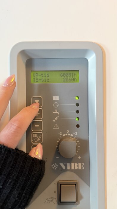 Kontrollpanel av en värmepump med en hand som trycker på en knapp. Displayen visar driftstider: VP-tid 60081h, TS-tid 2060h.