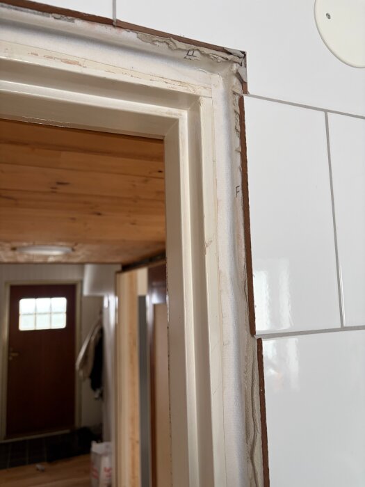 Närbild på en delvis borttagen dörrkarm i ett badrum med synlig tätskikt ovanför, omgiven av vita kakelplattor och trädetaljer.