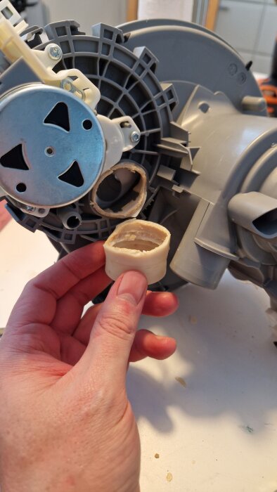 En hand håller ett avtagbart plastfilter framför en grå komponent med en synlig skruv och fläktblad.