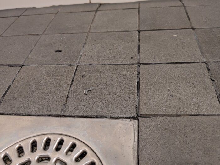 Spruckna fogar och lösa plattor på ett duschgolv med närbild av avloppsbrunn i ett badrum.