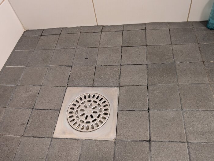 Närbild av grått kakelgolv i dusch med synliga sprickor i fogarna runt brunn.