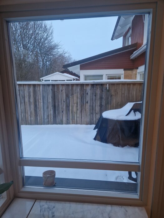 Utsikt genom ett radhusfönster, visar en snötäckt uteplats med en grill under ett överdrag och ett plank i bakgrunden.