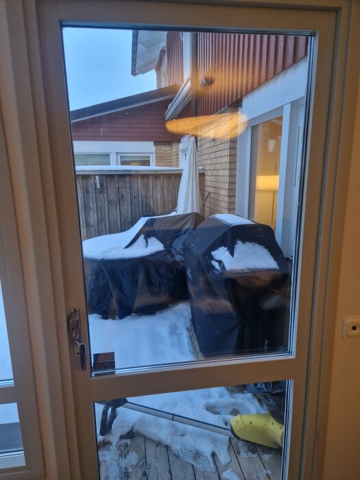 Vy genom fransk dörr ut mot snötäckt uteplats med täckta grillar och skyffel. Möjlig källa till kallt drag i radhus.