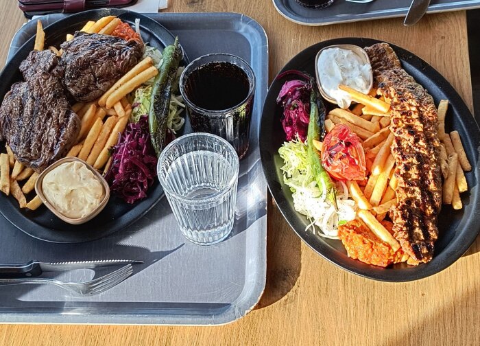 Två tallrikar med kött och pommes frites. Ena med entrecote, den andra med adanaspett, omgiven av sallad, sås och drycker.