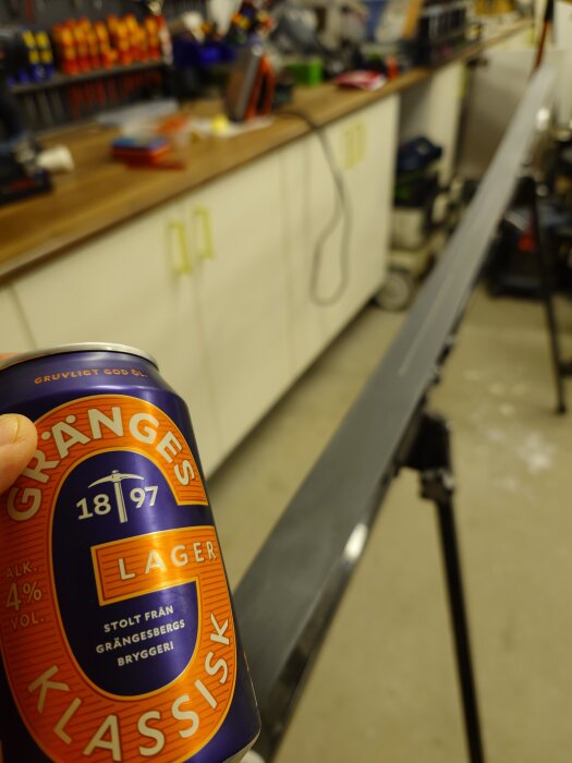 En hand håller en burk Gränges Klassisk lageröl framför en arbetsyta med skidvalla.