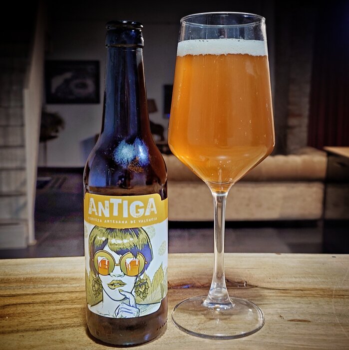 Flaska och glas med ljus öl märkt "ANTIGA". Bakgrund med soffa och tavla.