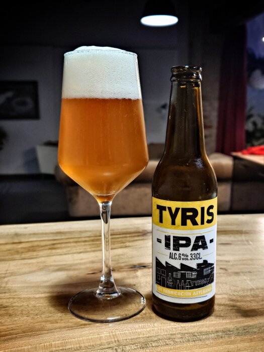 Ett glas med skummande öl bredvid en flaska Tyris IPA, 6% alkoholhalt, på ett träbord i en hemtrevlig miljö.