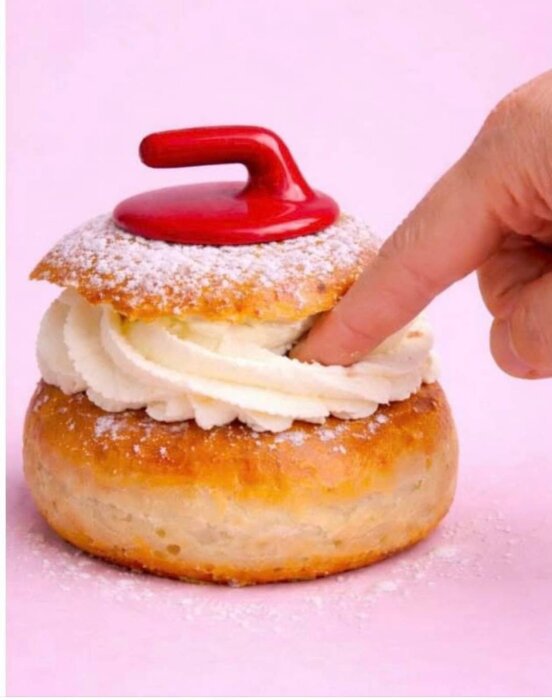 OS-semla med röd curlingsten på toppen, vispad grädde och bakelsebröd, finger trycker på grädden.