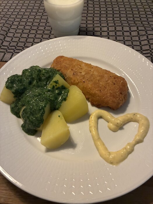 Fisk, potatis och spenat på en tallrik med en såsformad hjärta och ett glas mjölk i bakgrunden.