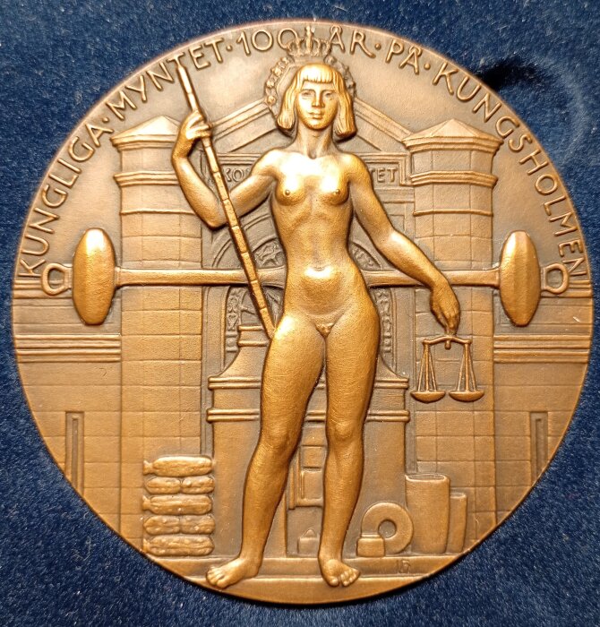 Medalj av Leo Holmgren med en kvinnlig figur som håller en stav och balansvåg, utförd med rika detaljer, texten "Kungliga Myntet 100 år på Kungsholmen".